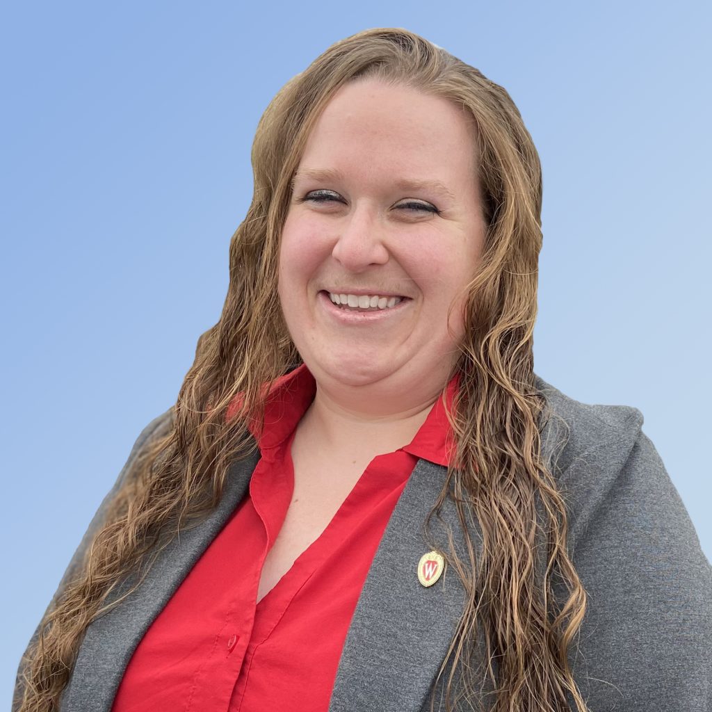 Head and shoulders shot of Lyssa Seefeldt.  Lyssa is wearing a red collared shirt and a gray blazer wtih a UW-Madison pin in the lapel.  She has long brown curly hair and is in front of a sky blue gradient background.