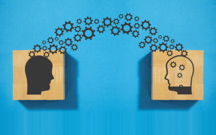 The image depicts two wooden silhouettes of human heads placed on a blue background. One head silhouette is solid black, while the other contains several small white gears or cogs inside it, suggesting the concept of a mind filled with thoughts, ideas, or machinery of thinking and cognitive processes. Small black gear shapes are arranged in an arched formation connecting the two head silhouettes, further reinforcing the metaphorical representation of the flow of thoughts or the mechanics of knowledge transfer between minds.