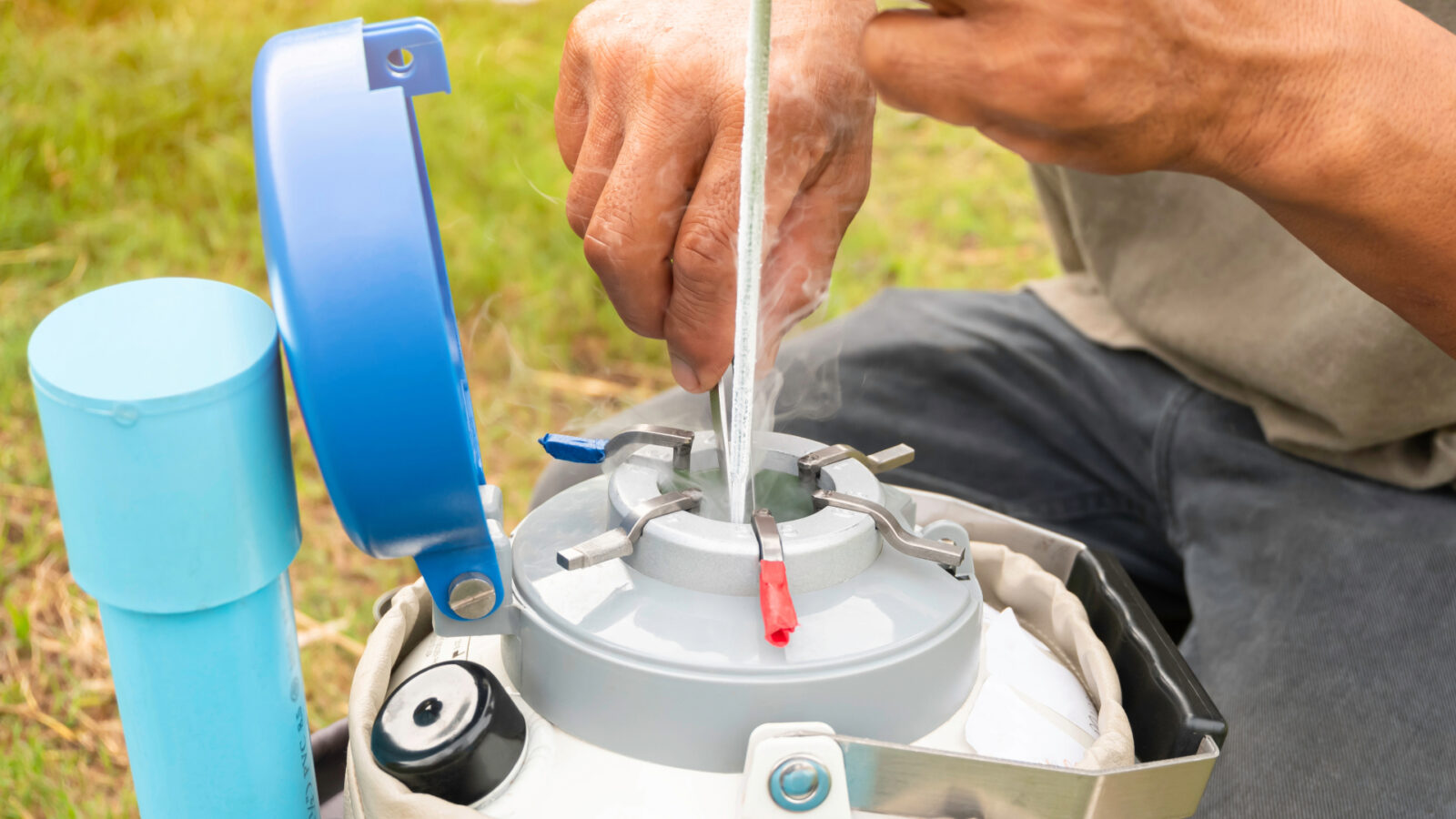 A person calibrating a scientific instrument in an outdoor field setting, pouring liquid from a plastic container into the device's chamber while holding measurement tools and components.