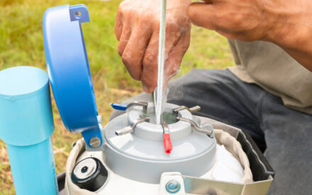 A person calibrating a scientific instrument in an outdoor field setting, pouring liquid from a plastic container into the device's chamber while holding measurement tools and components.