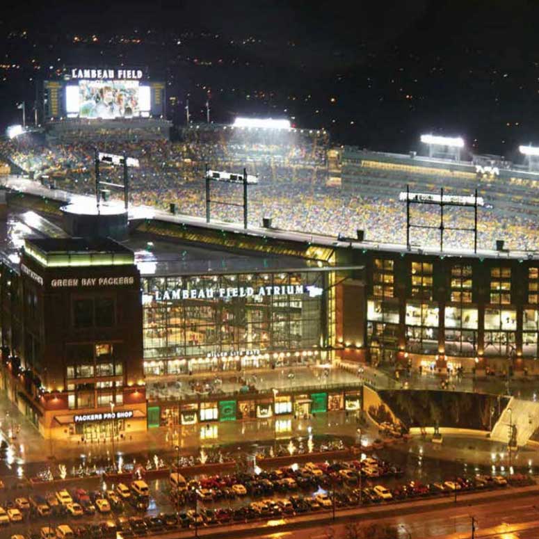 An aerial photograph of Lambeau Field in Green Bay, Wisconsin. It is night and the stadium is filled with fans. The exterior of the stadium shows cars parked in the parking lot.