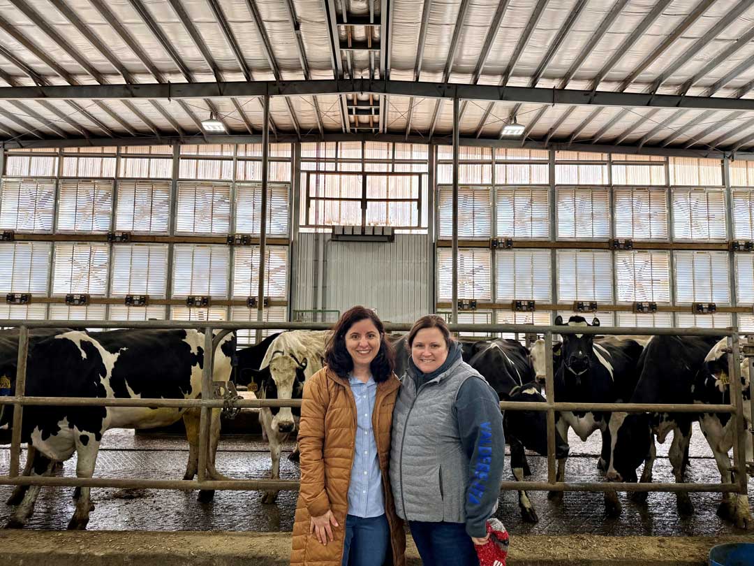 Two women stand in front of Holstein cattle in modern dairy barn with metal roof and ventilation systems