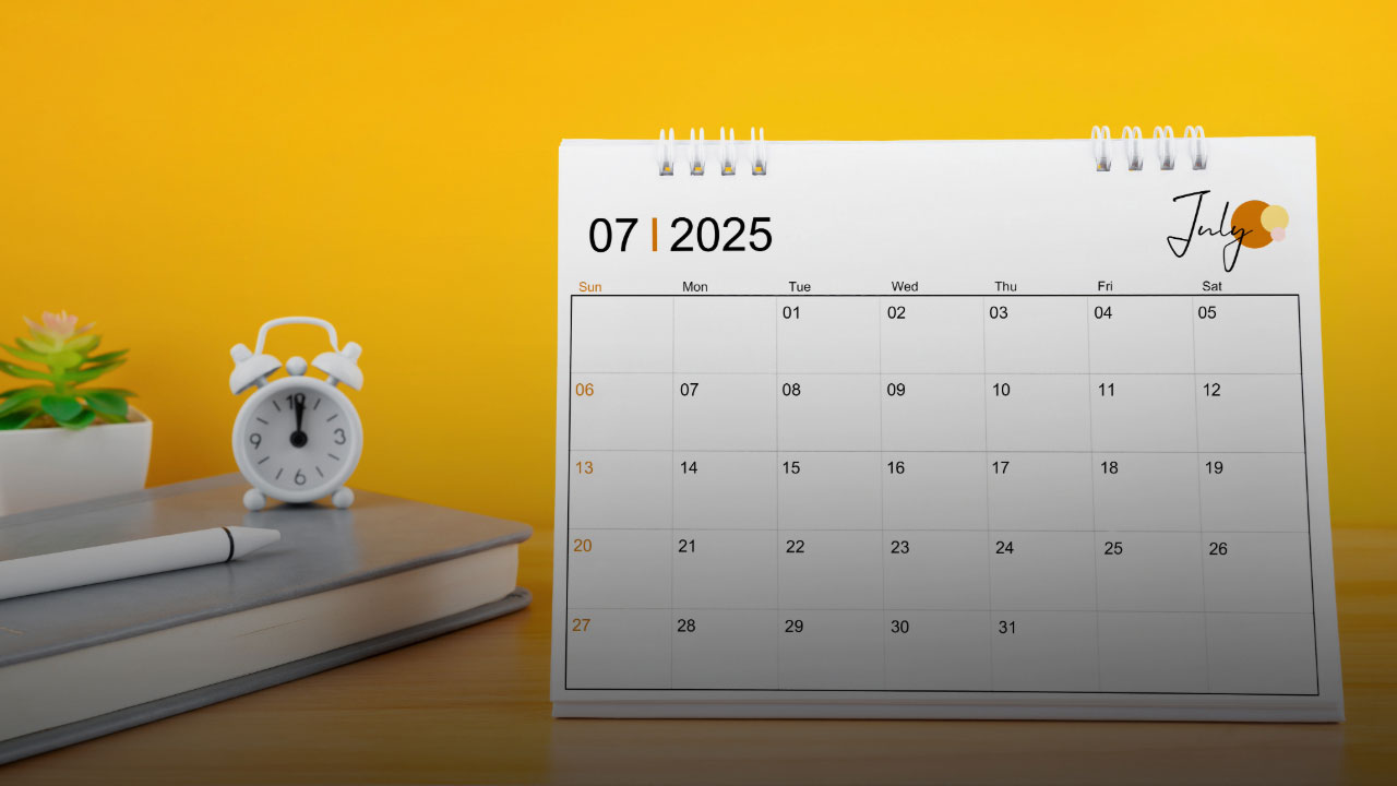July 2025 desk calendar showing monthly grid with dates 1-31, white alarm clock, notebook and pen on wooden desk