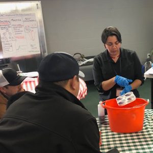A woman is demonstrating a procedure related to the dairy industry. She is wearing rubber gloves and holding 4 samples over a bucket while adult students observe.