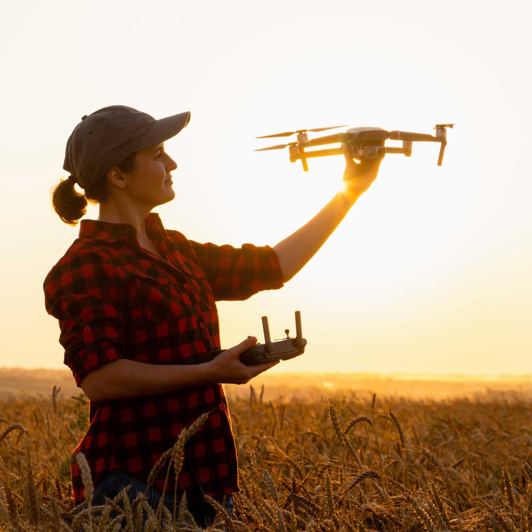A farmer stands in a field of wheat holding a flying drone above head level.