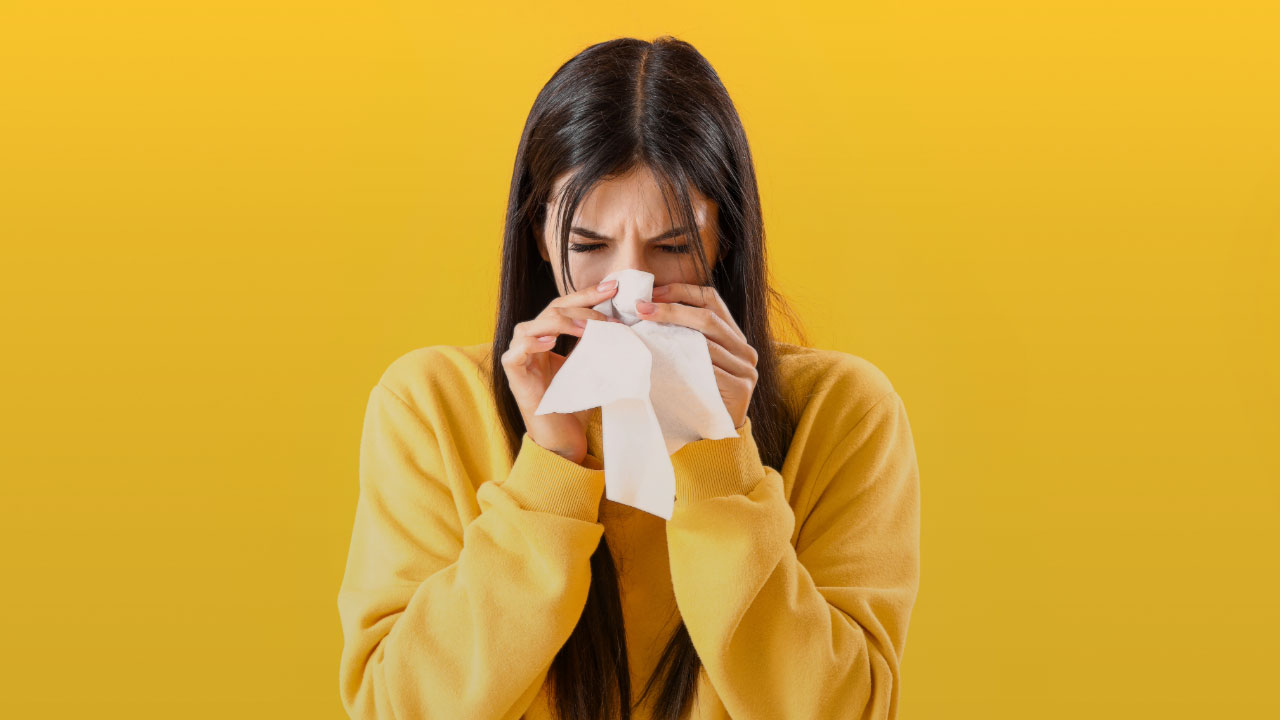 A woman holding a facial tissue up to her nose and covering her mouth. She's wearing a yellow sweater and the background is yellow.