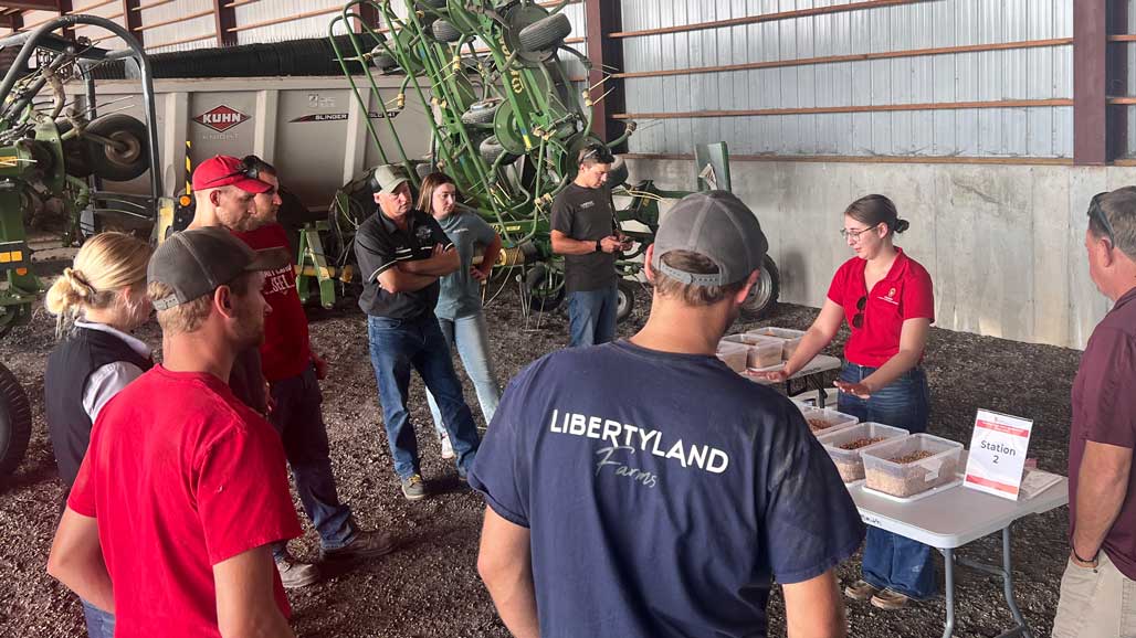 Extension educator shows soybean sample bins to farmers gathered inside a machine shed near a grain cart.